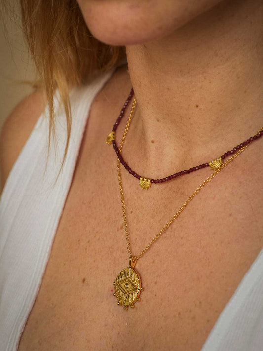 A close-up of a person wearing two layered necklaces: a gold-tone chain necklace with an evil eye protective pendant and a shorter dark red beaded necklace with small gold heart charms.