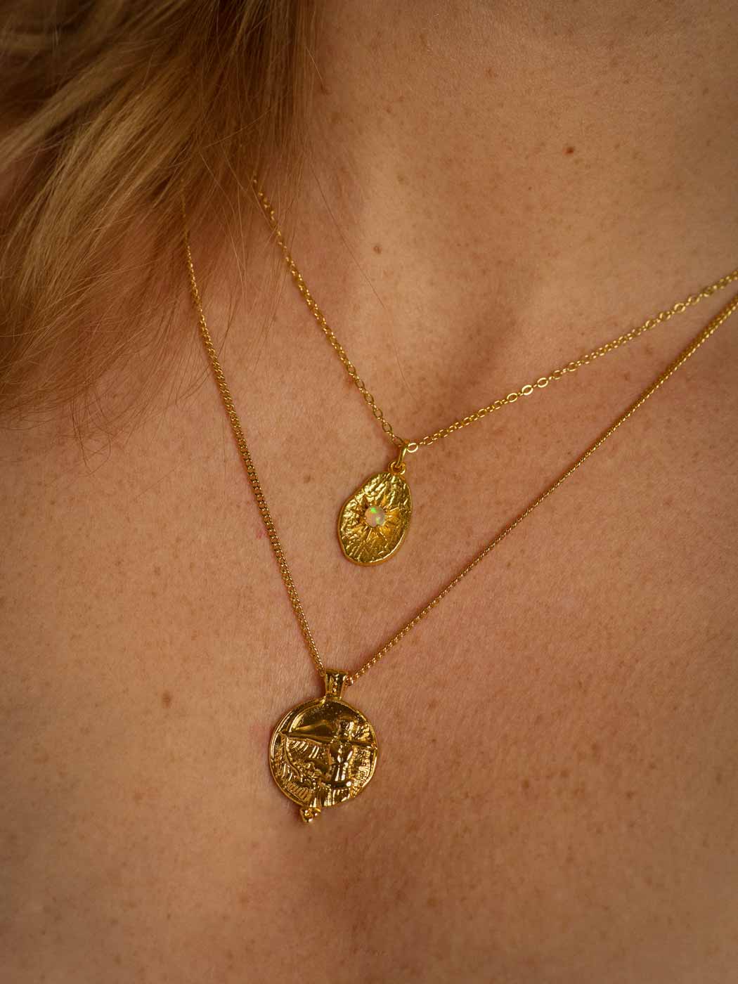 A close-up of a person's neck and upper chest wearing two layered gold necklaces. The shorter chain features a teardrop-shaped pendant with a opal at the center, while the longer chain holds a circular pendant with an embossed figure of goddess artemis