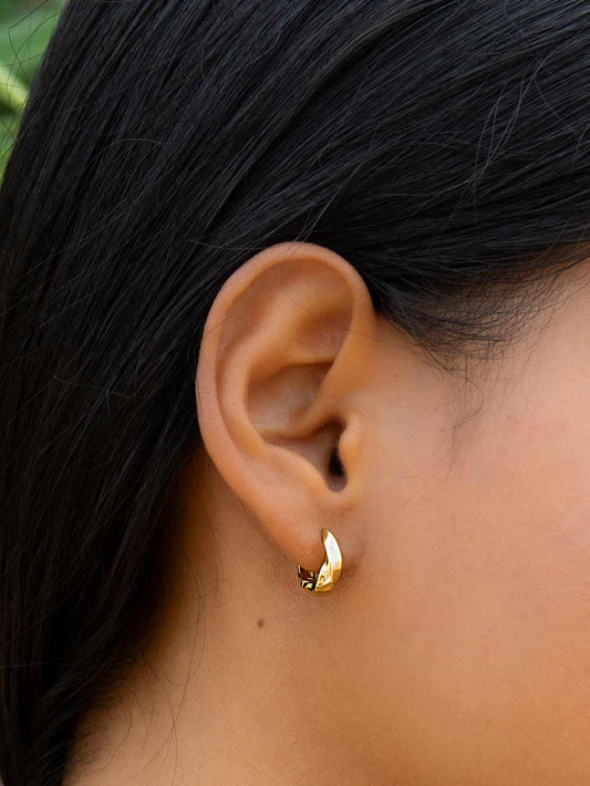 Close-up of an ear wearing a twisted gold huggie earring with dark hair and skin in the background