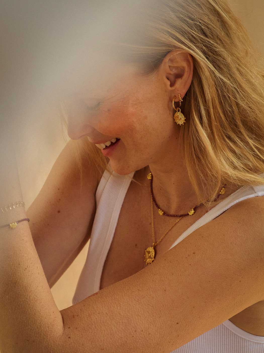 A close-up lifestyle shot of a woman wearing gold jewelry, including a gold dangle earring, layered gold necklaces, and a gold bracelet, paired with a white tank top.