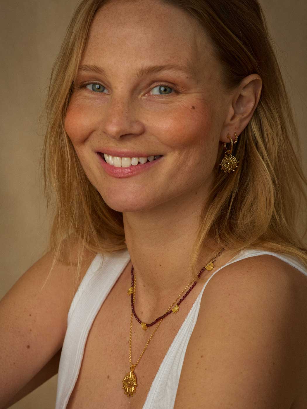 A smiling blonde woman wearing gold jewelry, including a gold pendant necklace, a beaded necklace, and a gold sunburst earring, against a warm beige background.