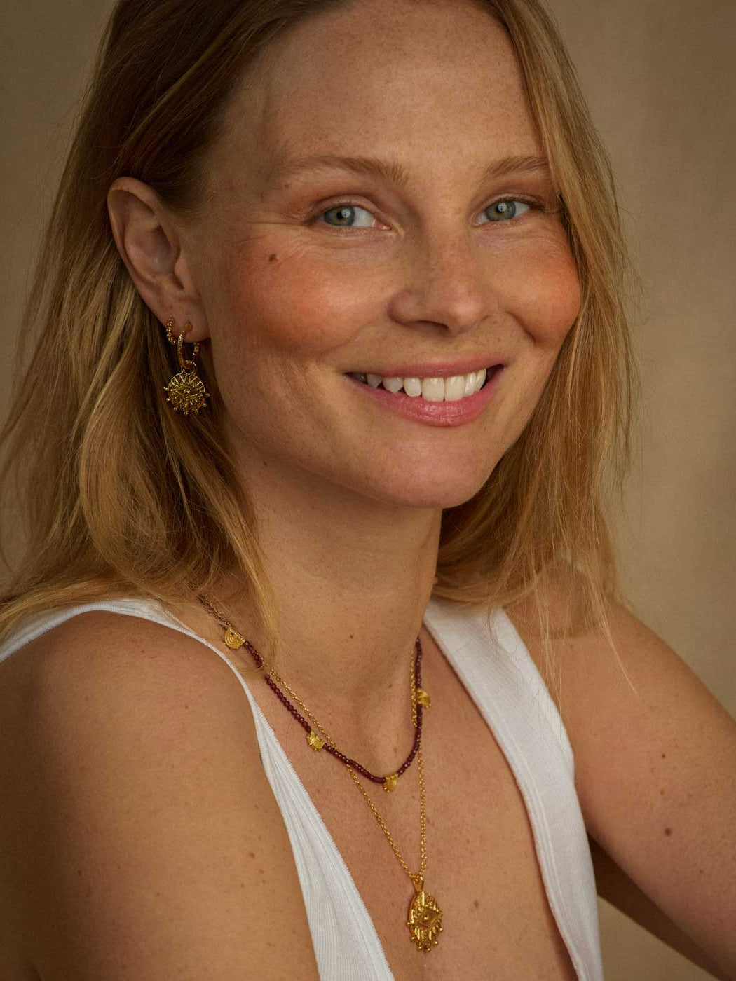 A smiling blonde woman wearing gold jewelry, including a gold pendant necklace, a beaded necklace, and a gold sunburst earring, against a warm beige background.