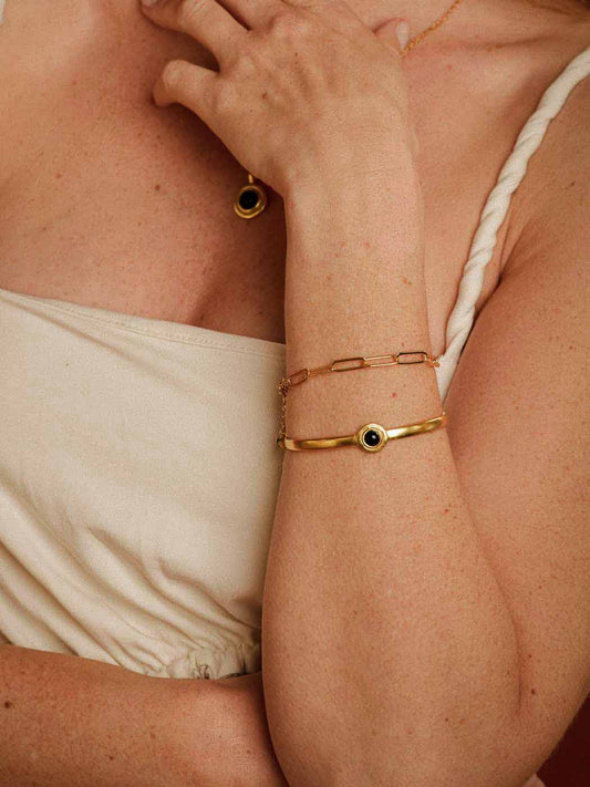 A close-up of a person's arm and chest wearing layered gold bracelets, including a bangle with a small black circular detail, alongside a matching gold drop earring.