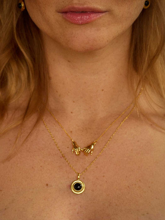 Close-up of a woman wearing layered gold necklaces, including a hand-holding pendant necklace and a circular pendant with a black agate center, on her neck and chest.