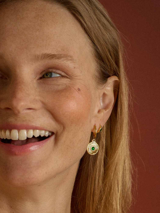 smiling woman wearing hoops that feature a round textured pendant and smaller central emerald gemstone