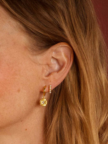 Close-up of a woman's ear wearing a small gold hoop earring with a dangling round gold charm and star diamante embossed in the center, and a slim vertical pearl stud earring.