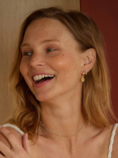 A close-up lifestyle shot of a smiling woman with blonde hair, wearing a delicate gold necklace and a small gold dangle earring, set against a warm neutral background.
