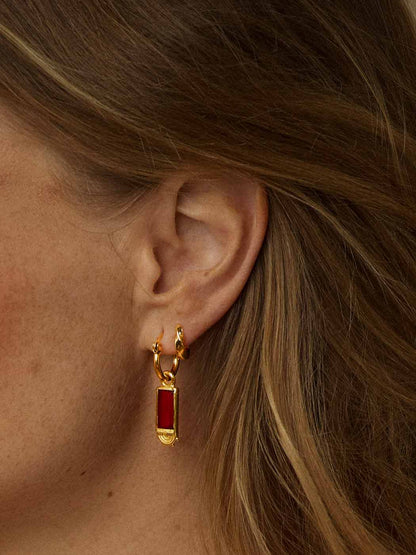 A close-up of a model's ear wearing a gold huggie hoop earring with a small rectangular red gemstone pendant.
