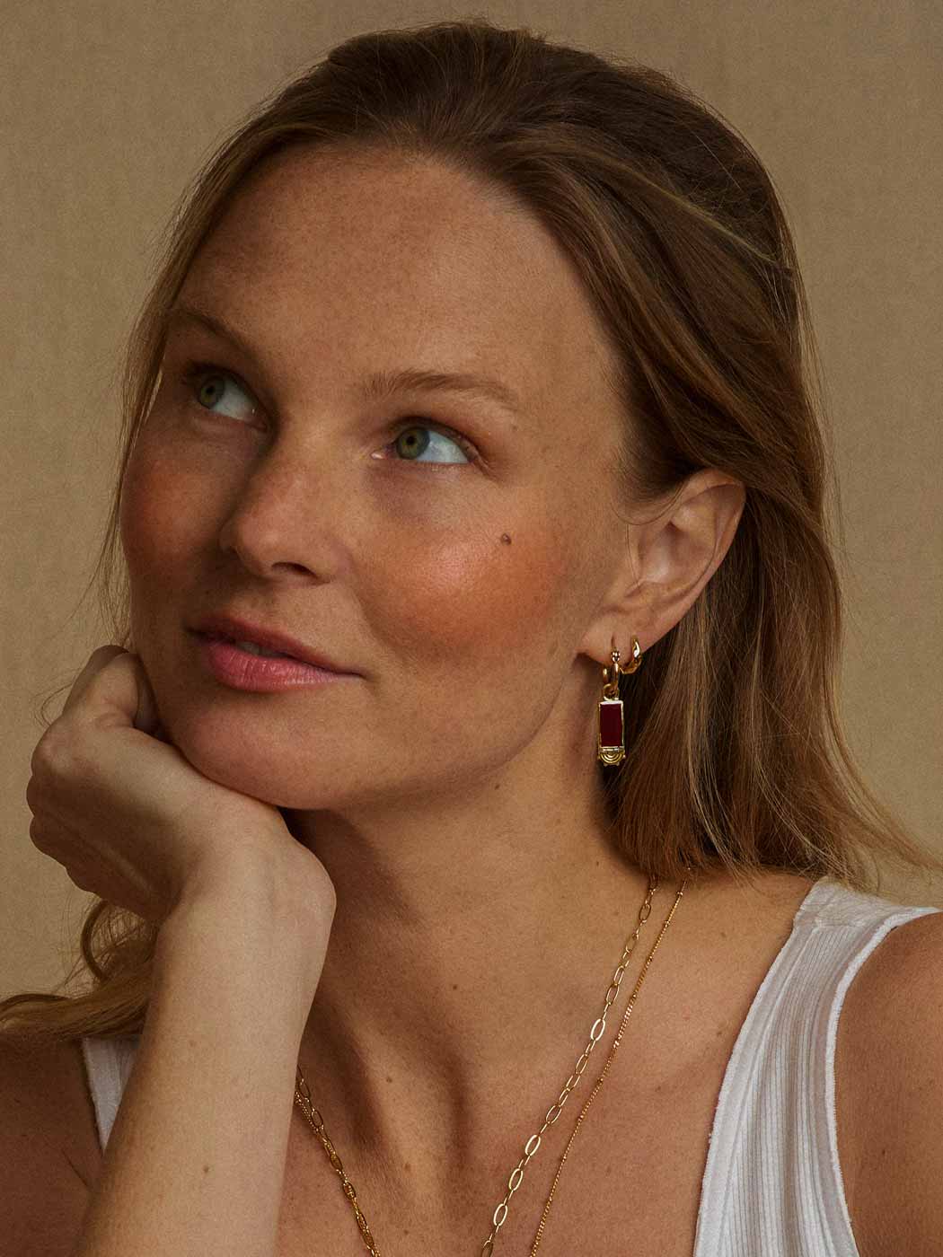 A close-up portrait of a woman with blonde hair, wearing a gold huggie hoop earring with a dark red rectangular drop pendant, layered with a delicate gold chain necklace, against a warm beige background.