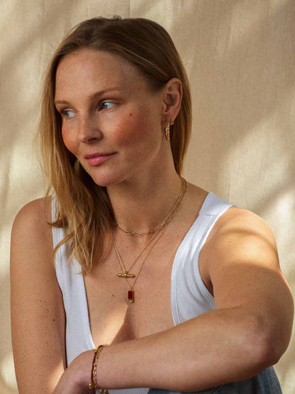 A woman wearing layered gold necklaces, one with a small red rectangular pendant, and gold bracelets, posing in a white tank top against a warm beige textured backdrop.