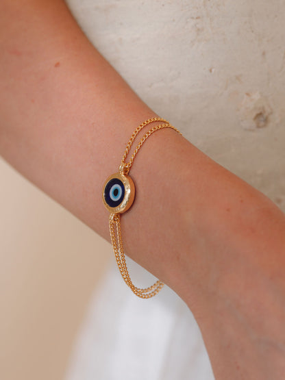 Gold bracelet with a blue eye charm on a wrist against a neutral background