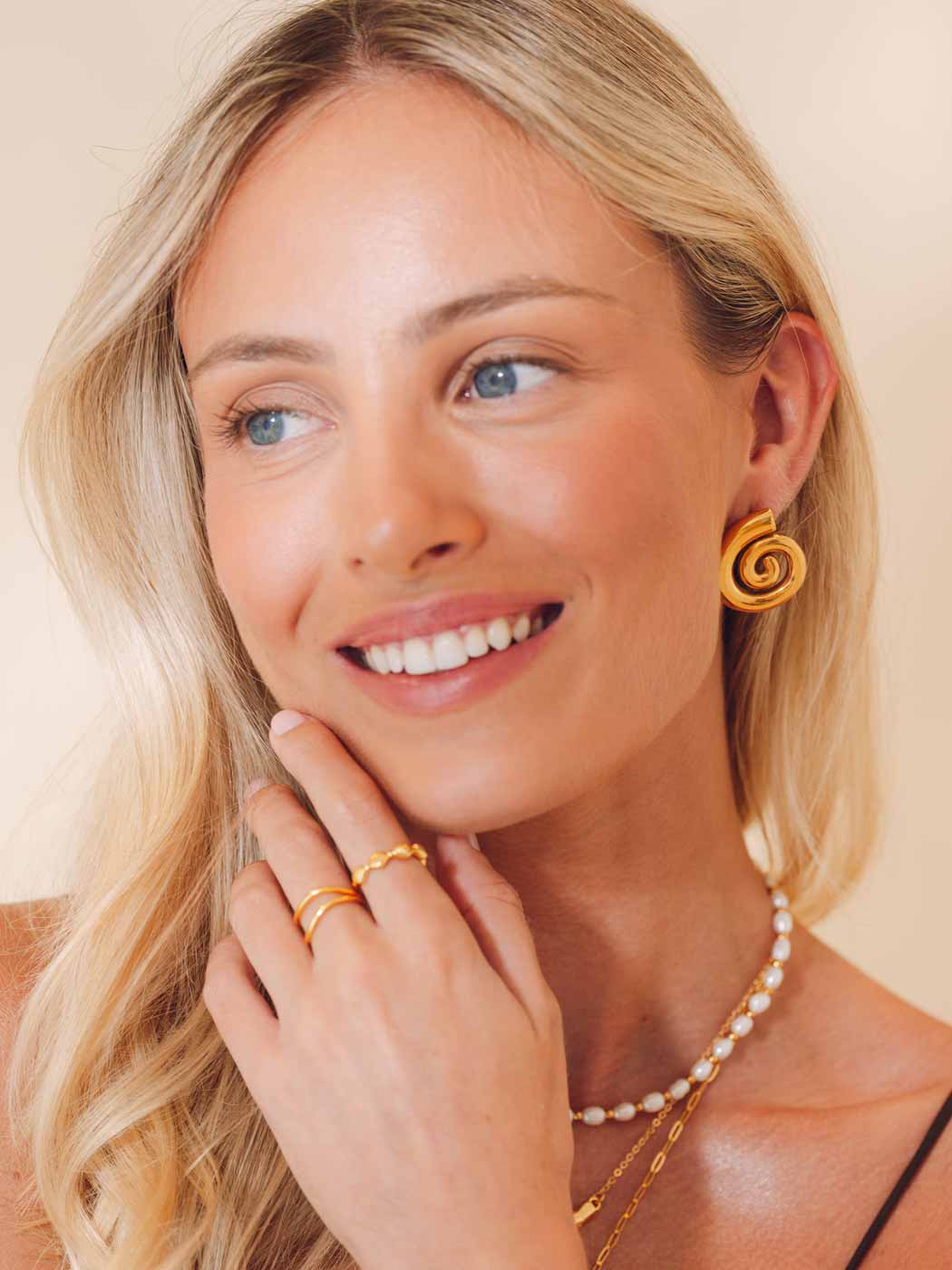 Woman wearing spiral gold earrings, rings, and a necklace on a beige background