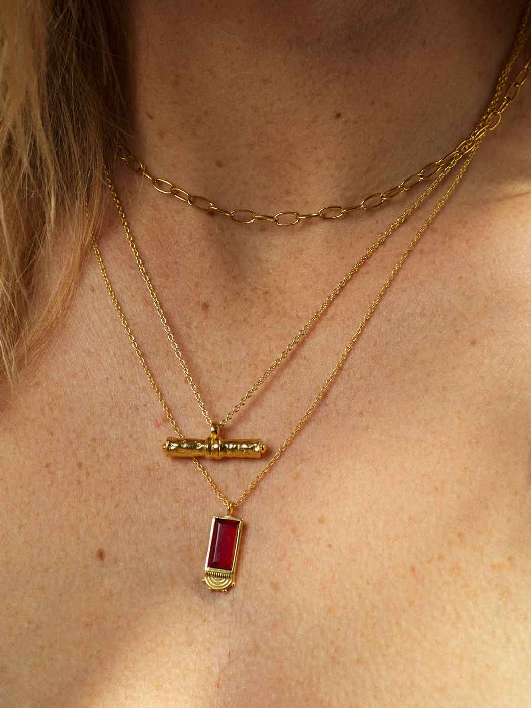 A close-up of a person's neck and chest wearing layered gold necklaces, including a curb chain, a delicate chain with a small gold t-bar pendant, and a longer chain with a rectangular red gemstone pendant.