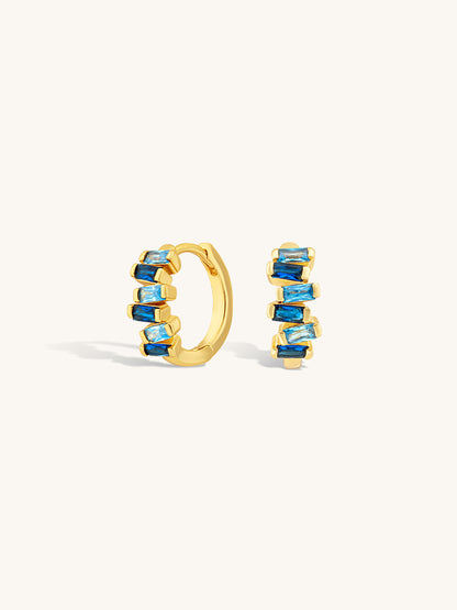Gold huggie earrings with sapphire blue gemstones on a white background