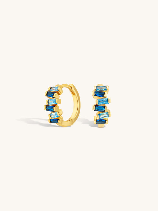Gold huggie earrings with sapphire blue gemstones on a white background