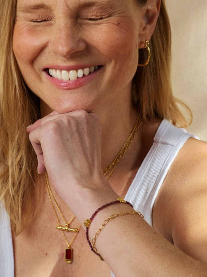 Close-up of a smiling woman wearing layered gold jewelry, including a necklace with a rectangular red gemstone pendant, a cross charm, and bracelets, along with a gold hoop earring.