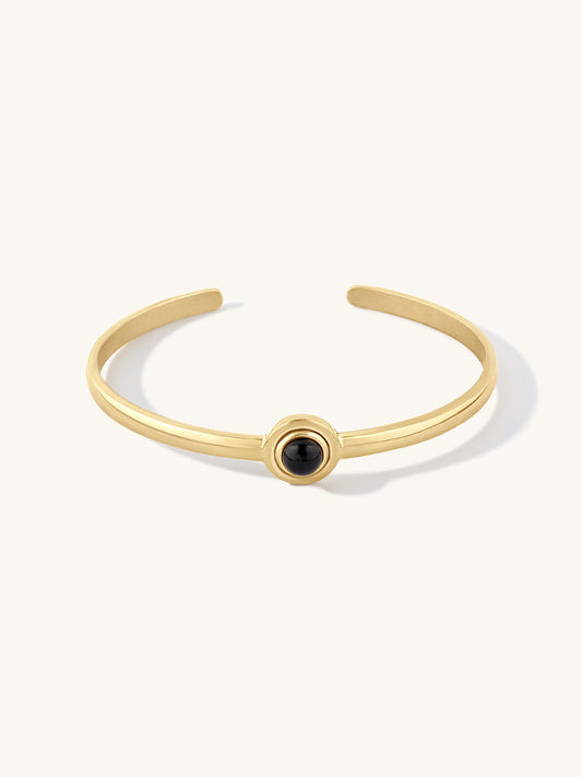 A slim gold cuff bracelet with a central circular bezel holding a small black agate stone, shown on a clean off-white background.