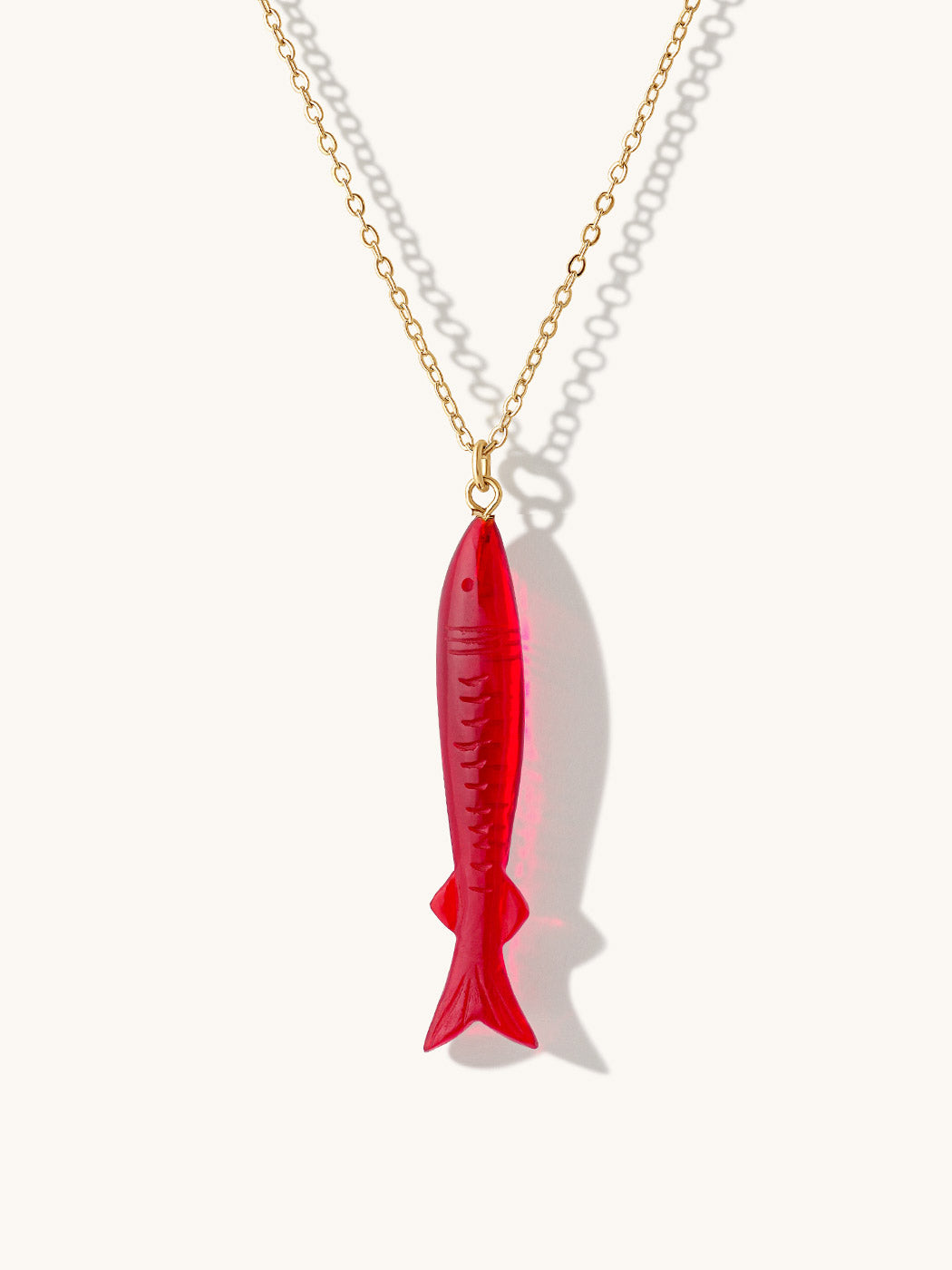 A gold chain necklace featuring a garnet red gemstone fish-shaped pendant, shown against a clean off-white background with soft shadows.