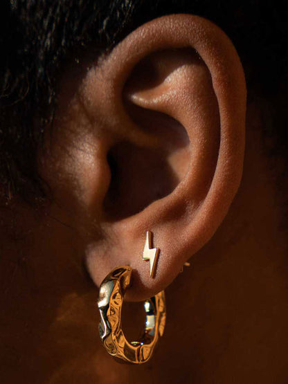A close-up of a person's ear showcasing a pair of gold earrings: a small lightning bolt stud earring and a textured, chunky hoop earring.