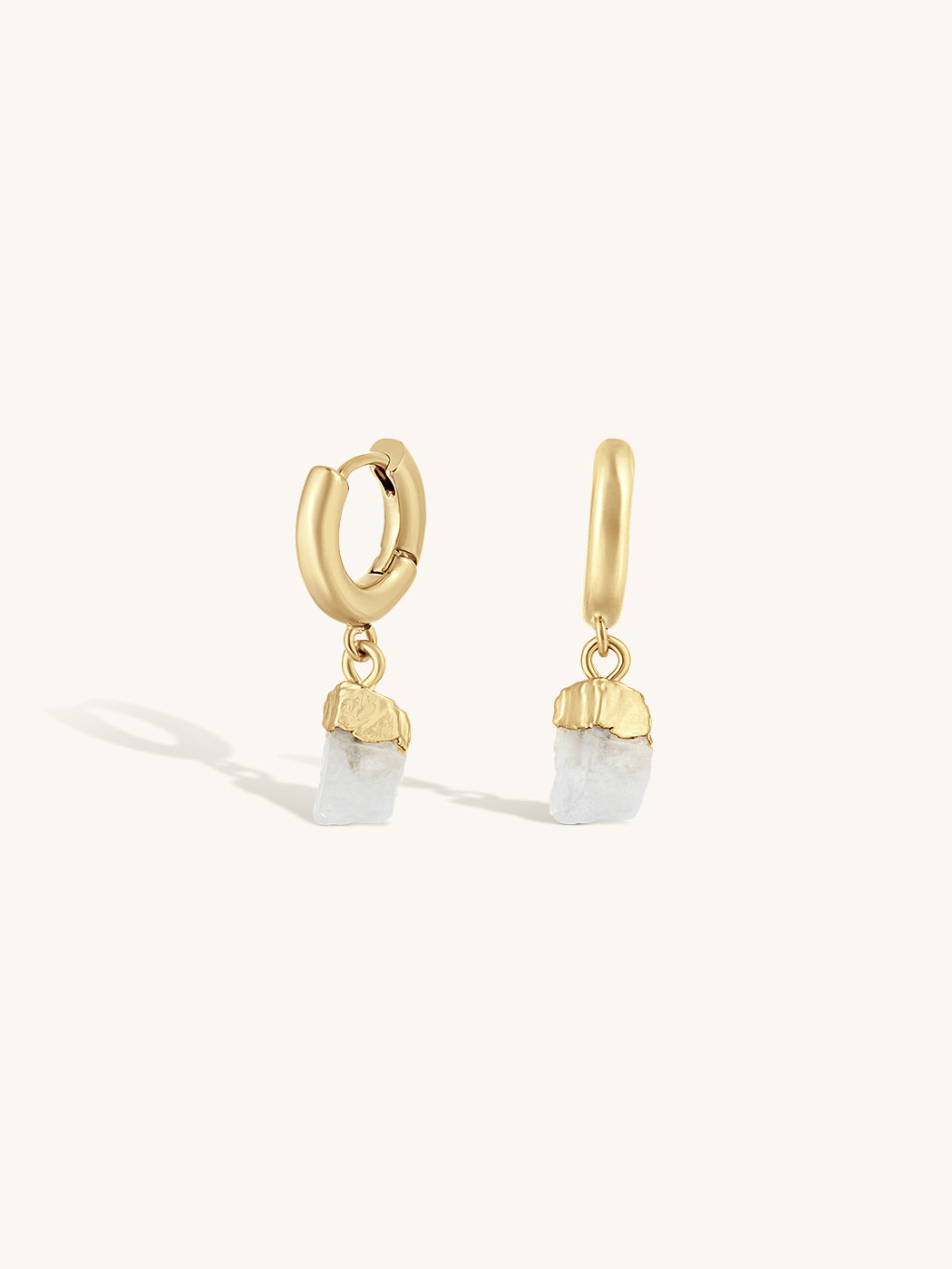 A pair of gold huggie hoop earrings with dangling raw, irregular-cut white gemstones, shown on a clean off-white background.