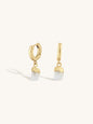 A pair of gold huggie hoop earrings with dangling raw, irregular-cut white gemstones, shown on a clean off-white background.