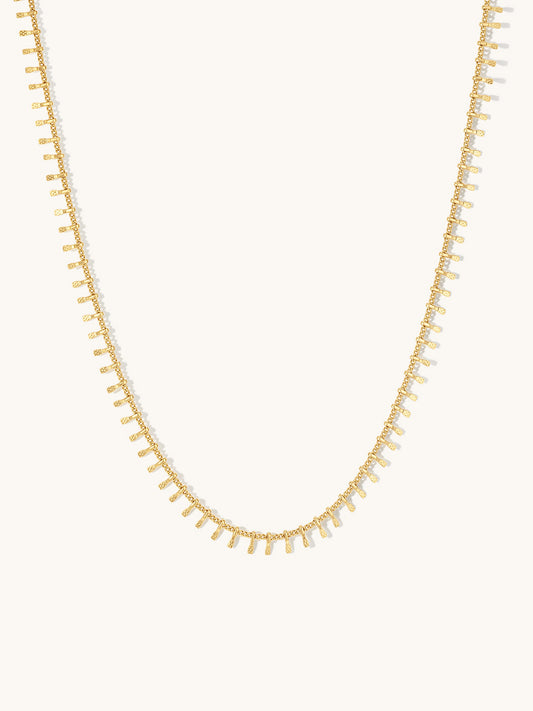 A delicate gold chain necklace featuring small, regularly spaced rectangular drop charms, displayed against a solid off-white background.