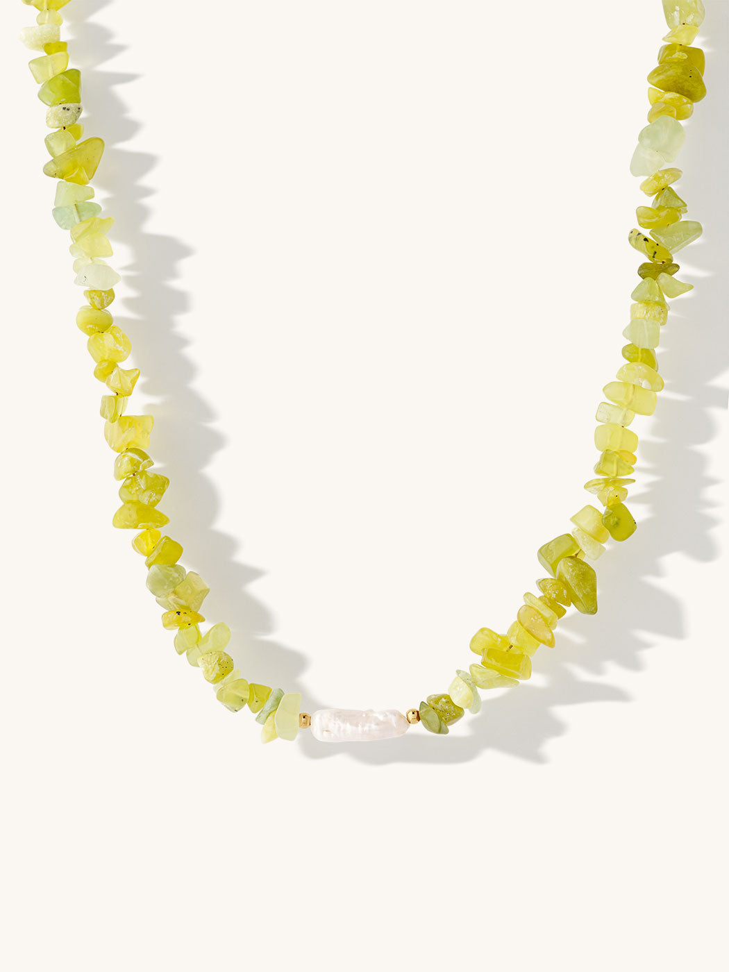 A beaded necklace made of irregular yellow-green peridot gemstone chips with a freshwater pearl and gold spacer, displayed on a light cream background with soft shadows.