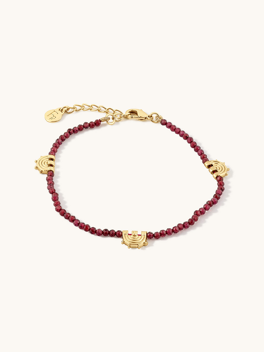 A delicate gold chain bracelet featuring small, faceted garnet beads and three gold carved charms, displayed on a clean off-white background.